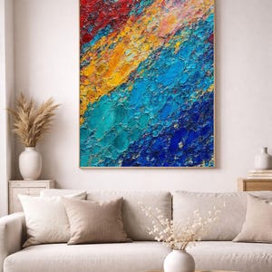 Op de afbeelding: Abstract textuurschilderij met rode, gele en blauwe tinten in een gouden lijst. Het hangt boven een beige bank met neutrale kussens. Een ronde houten salontafel staat voor de bank.