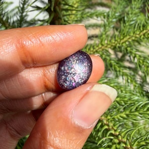 May include: A small, oval-shaped gemstone with a dark purple base and iridescent flecks of silver, pink, and blue. The stone is held between fingers against a backdrop of green foliage. The gemstone's surface reflects light, creating a shimmering effect.