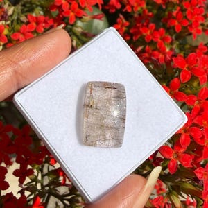 Pode incluir: Uma pedra preciosa retangular com uma base transparente e inclusões douradas e pretas, apresentada numa caixa branca. A pedra é exibida contra um fundo de vibrantes flores vermelhas. A superfície da pedra reflete a luz, realçando os seus padrões únicos.