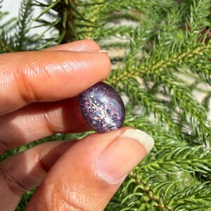 May include: A dark purple, oval-shaped gemstone with iridescent flecks of color. The stone is held between fingers against a backdrop of green foliage. The gemstone's surface reflects light, showcasing a mix of pink, silver, and blue hues.