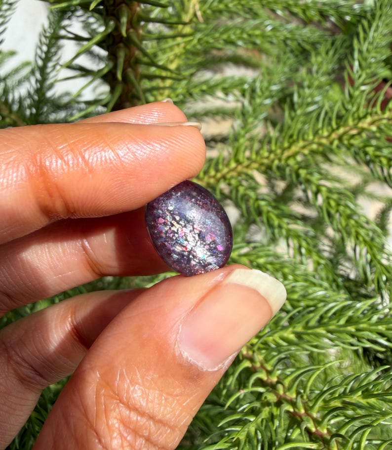 May include: A dark purple, oval-shaped gemstone with iridescent flecks of color. The stone is held between fingers against a backdrop of green foliage. The gemstone's surface reflects light, showcasing a mix of pink, silver, and blue hues.
