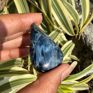 Peut inclure: Une pierre précieuse polie en forme de goutte d'eau, d'un bleu profond avec des motifs tourbillonnants blancs et dorés. La pierre est tenue devant un fond de feuillage vert et jaune, soulignant sa beauté naturelle.