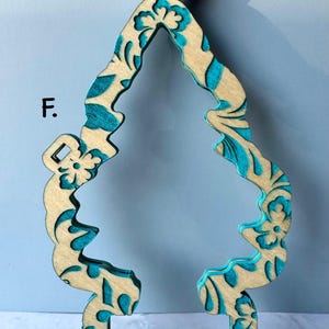 May include: Wooden ornament in the shape of a stylized Christmas tree. The ornament has a natural wood color with teal blue floral patterns and accents. The ornament has a loop at the top for hanging.