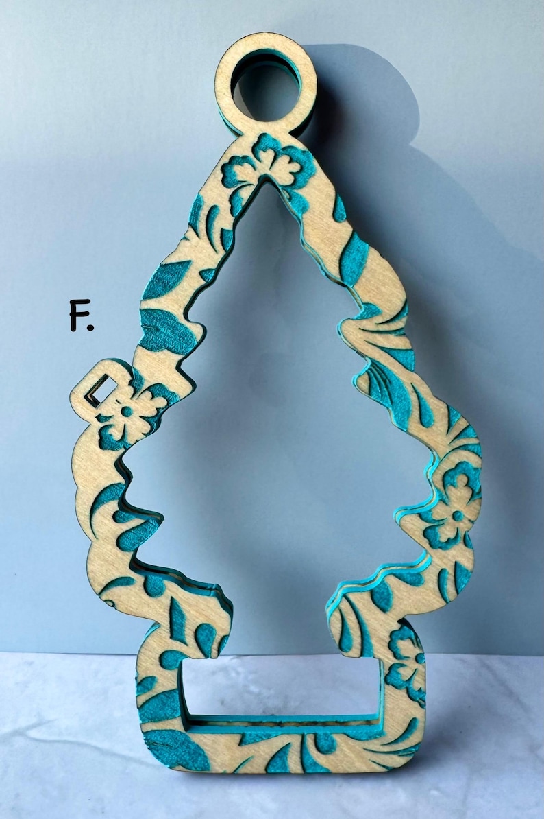 May include: Wooden ornament in the shape of a stylized Christmas tree. The ornament has a natural wood color with teal blue floral patterns and accents. The ornament has a loop at the top for hanging.