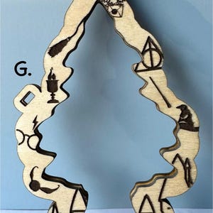 May include: Wooden ornament in the shape of a stylized Christmas tree. The ornament is etched with various symbols, including an owl, lightning bolt, glasses, and a castle. The ornament has a hole at the top for hanging.