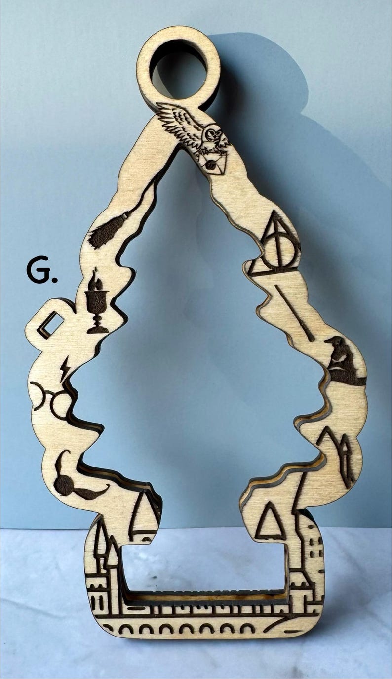 May include: Wooden ornament in the shape of a stylized Christmas tree. The ornament is etched with various symbols, including an owl, lightning bolt, glasses, and a castle. The ornament has a hole at the top for hanging.