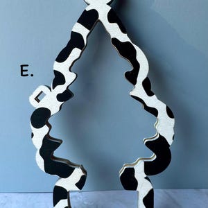 May include: Wooden ornament in the shape of a Christmas tree with a black and white cow print pattern. The ornament has a hole at the top for hanging and a rectangular base. The letter "E" is visible on the left side.