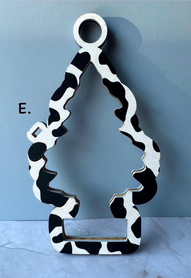 May include: Wooden ornament in the shape of a Christmas tree with a black and white cow print pattern. The ornament has a hole at the top for hanging and a rectangular base. The letter "E" is visible on the left side.