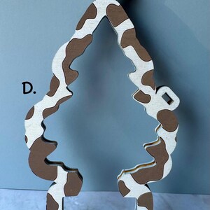 May include: A wooden ornament in the shape of a Christmas tree with a cow print pattern in brown and white. The ornament has a loop at the top for hanging and a rectangular base.