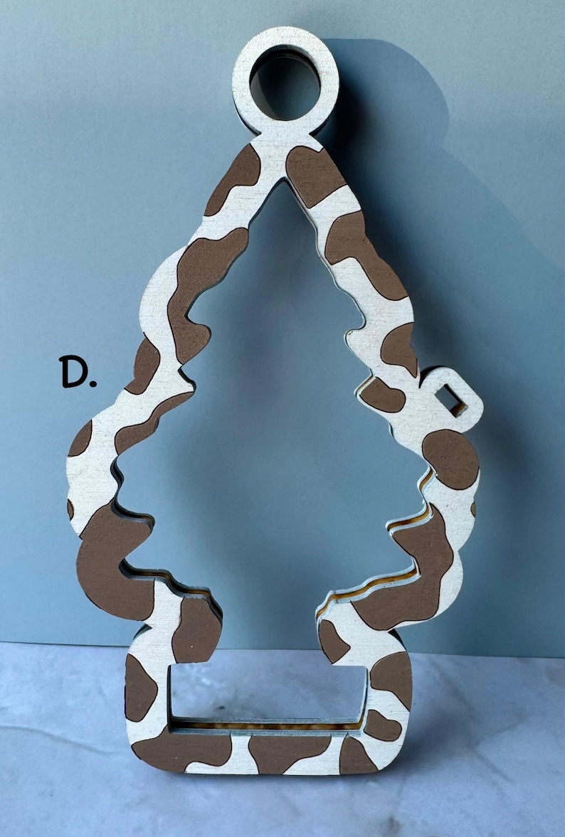 May include: A wooden ornament in the shape of a Christmas tree with a cow print pattern in brown and white. The ornament has a loop at the top for hanging and a rectangular base.