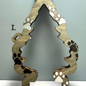 May include: Wooden Christmas tree ornament with paw print design. The ornament is shaped like a tree with a circular hole at the top for hanging. The paw prints are in black and white. The ornament is made of wood.