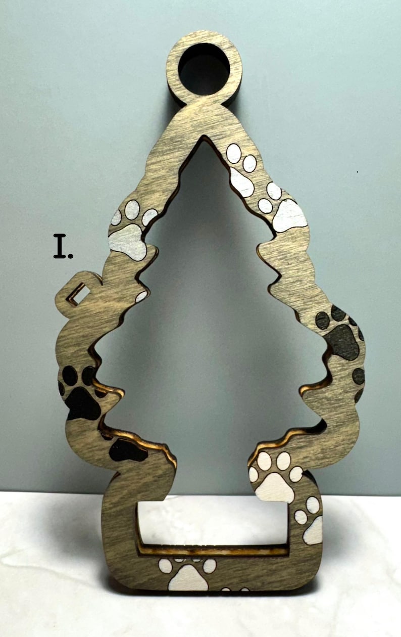 May include: Wooden Christmas tree ornament with paw print design. The ornament is shaped like a tree with a circular hole at the top for hanging. The paw prints are in black and white. The ornament is made of wood.