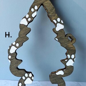 May include: Wooden Christmas tree ornament in a brown finish, shaped like a tree with a loop at the top for hanging. The tree is decorated with white paw prints. The letter "H" is visible on the left side.