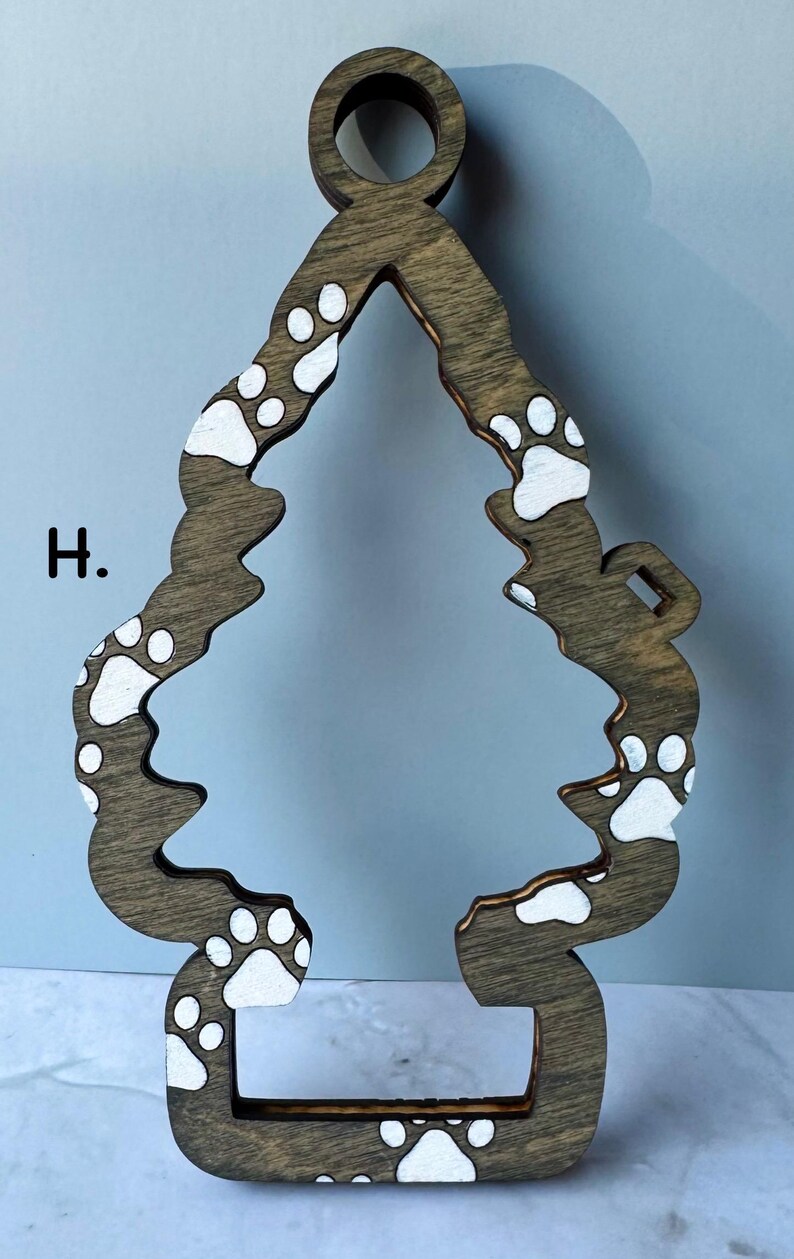 May include: Wooden Christmas tree ornament in a brown finish, shaped like a tree with a loop at the top for hanging. The tree is decorated with white paw prints. The letter "H" is visible on the left side.