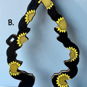 May include: Black wooden air freshener with a sunflower design. The air freshener is shaped like a stylized tree, with yellow sunflowers painted on a black background. The top has a circular hole for hanging.