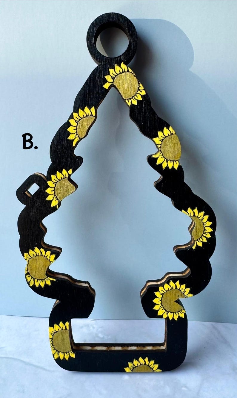 May include: Black wooden air freshener with a sunflower design. The air freshener is shaped like a stylized tree, with yellow sunflowers painted on a black background. The top has a circular hole for hanging.