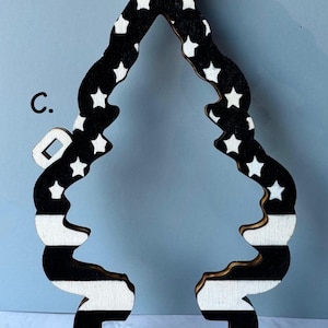 May include: Wooden Christmas tree ornament with a black and white design. The top has a circular opening for hanging. The tree shape features white stars on a black background and black and white stripes.