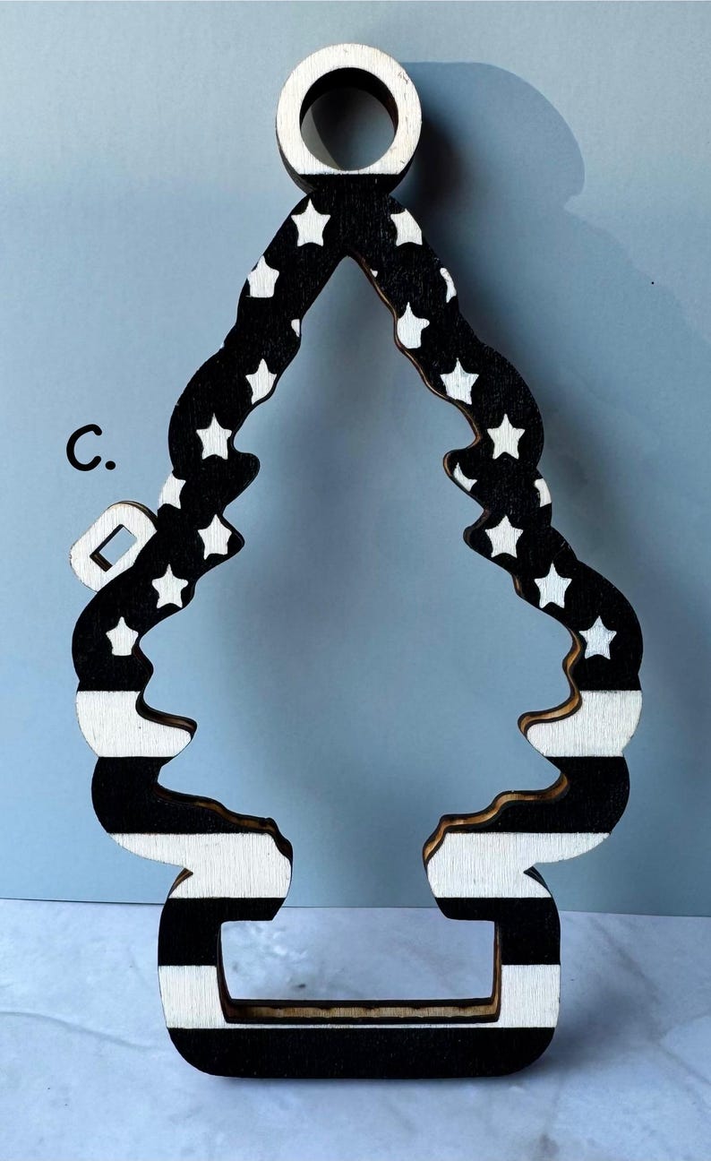 May include: Wooden Christmas tree ornament with a black and white design. The top has a circular opening for hanging. The tree shape features white stars on a black background and black and white stripes.