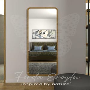Puede incluir: Un espejo rectangular de cuerpo entero con un marco de madera marrón claro. El espejo refleja una escena de dormitorio con una cama, obras de arte y una alfombra gris. El texto "Ferda Eroğlu inspired by nature" está en la parte inferior de la imagen.