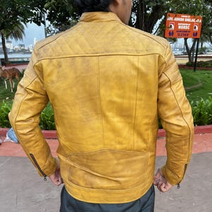 K&ouml;nnte beinhalten: Senfgelbe Lederjacke mit Steppung an den Schultern und Rei&szlig;verschl&uuml;ssen an den &Auml;rmeln. Die Jacke hat einen klassischen Biker-Stil mit Stehkragen und taillierter Passform. Die R&uuml;ckansicht zeigt die Details der Jacke, einschlie&szlig;lich der N&auml;hte.