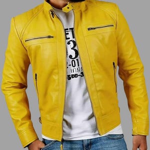 Op de afbeelding: Een felgele leren jas met een opstaande kraag en meerdere zakken met ritssluiting. De jas heeft een ritssluiting aan de voorkant en wordt gedragen over een wit t-shirt met zwarte tekst. Het model draagt een blauwe jeans.