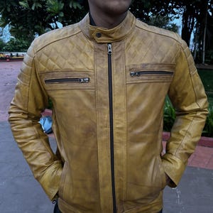 K&ouml;nnte beinhalten: Senffarbene Lederjacke mit Steppdesign an den Schultern und zwei Brusttaschen mit Rei&szlig;verschluss. Die Jacke hat einen Stehkragen und einen durchgehenden Rei&szlig;verschluss. Das Model tr&auml;gt die Jacke mit den H&auml;nden in den Taschen.