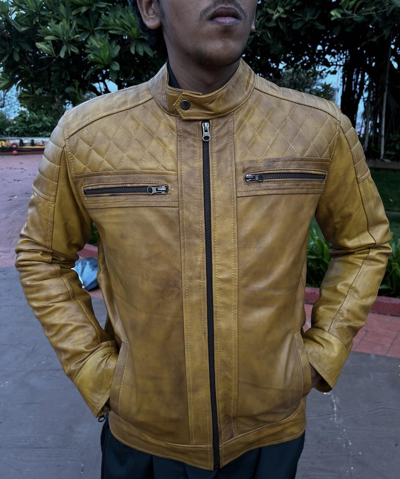 K&ouml;nnte beinhalten: Senffarbene Lederjacke mit Steppdesign an den Schultern und zwei Brusttaschen mit Rei&szlig;verschluss. Die Jacke hat einen Stehkragen und einen durchgehenden Rei&szlig;verschluss. Das Model tr&auml;gt die Jacke mit den H&auml;nden in den Taschen.