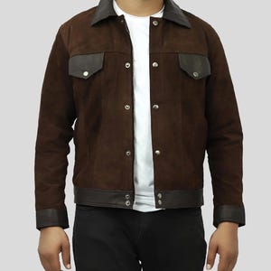 Puede incluir: Chaqueta de ante marrón con cuello, detalles de bolsillo y ribetes de cuero marrón oscuro. La chaqueta tiene cierre de botones y dos bolsillos en el pecho. El modelo lleva una camiseta blanca y pantalones negros.