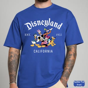 May include: Royal blue t-shirt featuring the Disneyland logo with Mickey Mouse, Minnie Mouse, Donald Duck, Goofy, Pluto, and Daisy Duck. The shirt includes the text "EST. 1955 CALIFORNIA".