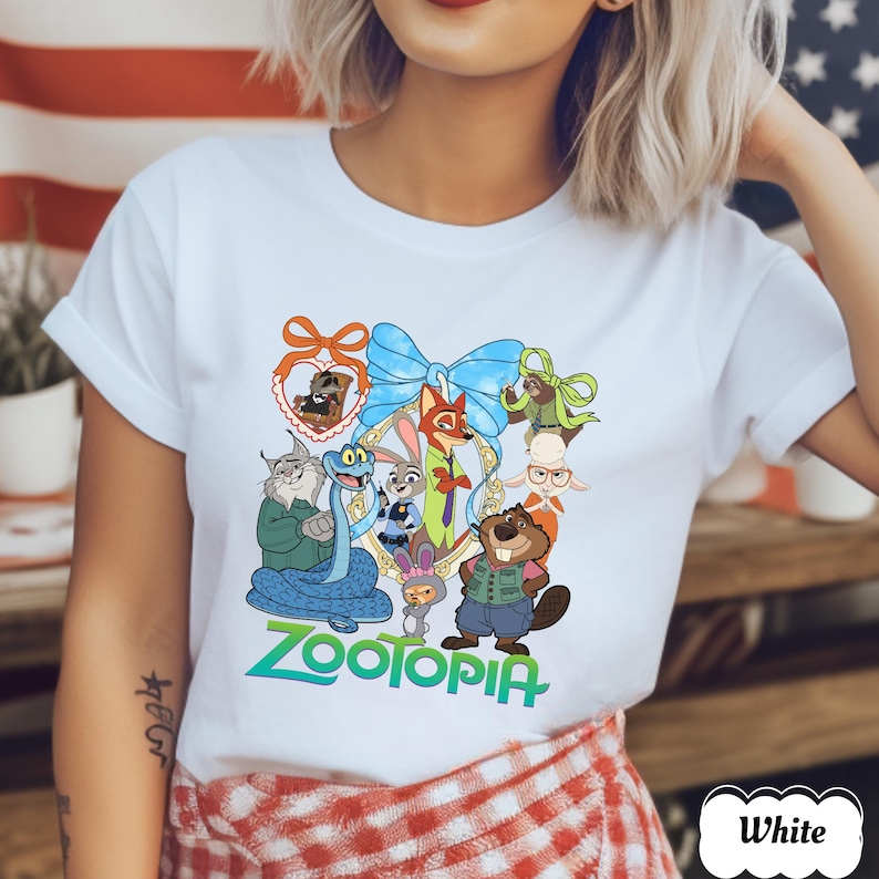 Może przedstawiać: Biała koszulka z kolorową grafiką postaci z filmu Zootopia. Projekt zawiera niebieską kokardę, złotą ramkę oraz napis "Zootopia" w kolorze zielonym i niebieskim. Koszulka wykonana jest z miękkiego materiału.