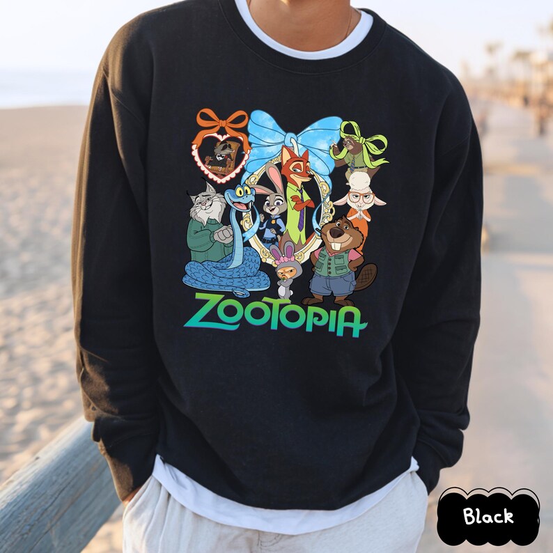 K&ouml;nnte beinhalten: Schwarzes Sweatshirt mit einer farbenfrohen Grafik von Charakteren aus dem Film "Zoomania". Das Design beinhaltet eine blaue Schleife, einen herzf&ouml;rmigen Rahmen und das Wort "Zootopia" in gr&uuml;nen und blauen Buchstaben.