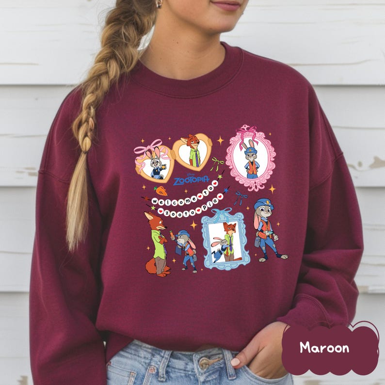 May include: Maroon sweatshirt featuring Zootopia characters in framed illustrations. The design includes images of a fox and a rabbit, with the word "Zootopia" in the center. The sweatshirt is a solid maroon color.