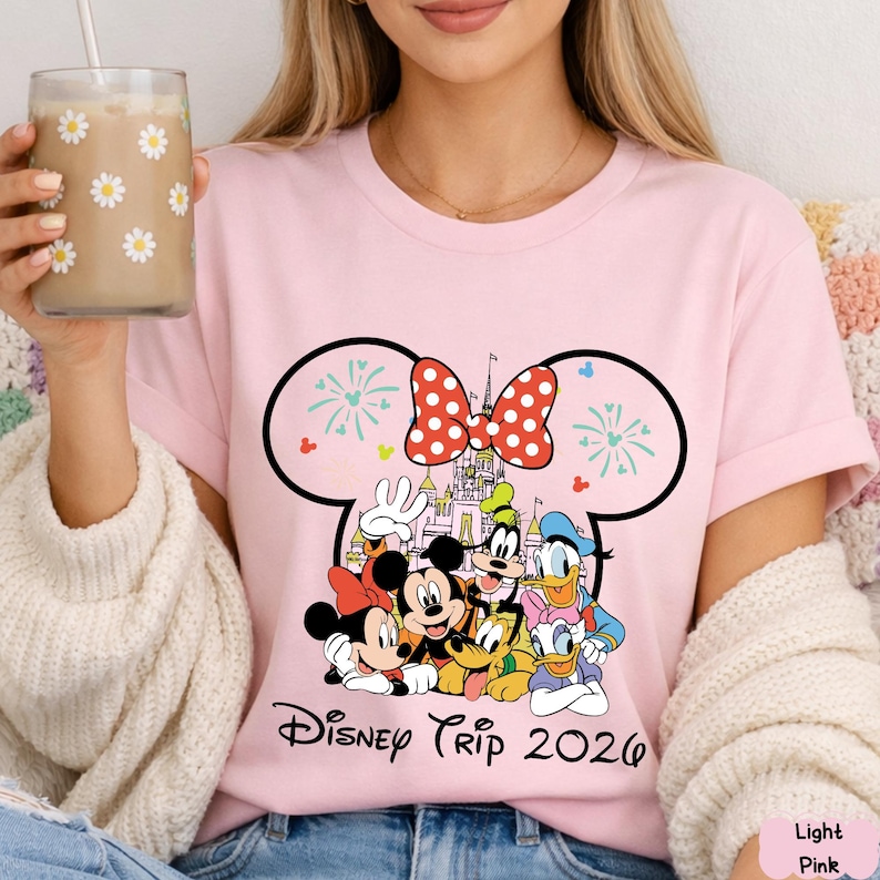 May include: Light pink t-shirt featuring a Disney Trip 2026 design. The graphic includes Mickey and Minnie Mouse, Goofy, Donald Duck, and Pluto, with a castle and fireworks. The design is framed by a black outline of Mickey Mouse ears.