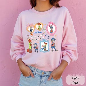 May include: Light pink sweatshirt featuring a colorful graphic of characters from the animated film "Zootopia." The design includes framed images of the characters, the film's title, and the phrase "Welcome to Zootopia."