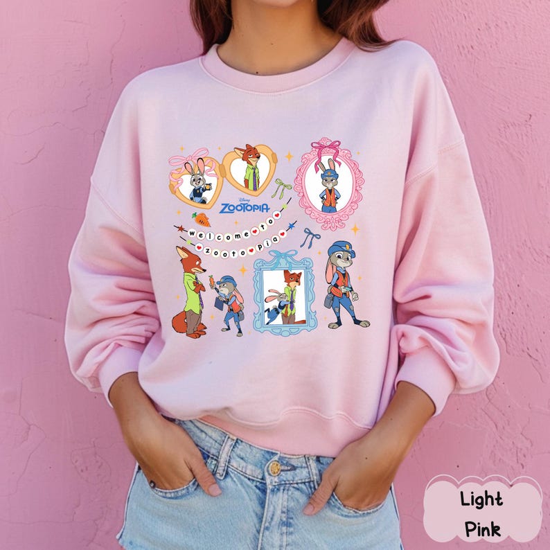 May include: Light pink sweatshirt featuring a colorful graphic of characters from the animated film "Zootopia." The design includes framed images of the characters, the film's title, and the phrase "Welcome to Zootopia."