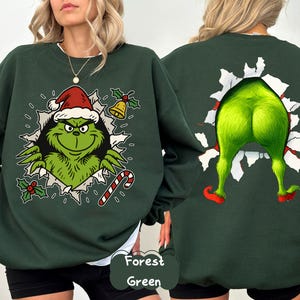 Puede incluir: Sudadera verde bosque con un gráfico del Grinch en la parte delantera, con un gorro de Papá Noel y un bastón de caramelo. La parte trasera de la sudadera muestra el trasero y las piernas verdes del Grinch. El texto "Forest Green" está en la parte inferior derecha.
