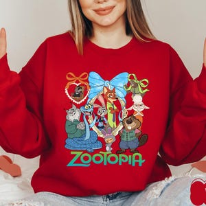 K&ouml;nnte beinhalten: Ein rotes Sweatshirt mit einer farbenfrohen Grafik von Charakteren aus dem Animationsfilm Zootopia. Das Design umfasst Judy Hopps, Nick Wilde und andere Charaktere, mit dem Wort "Zootopia" in gr&uuml;nen Buchstaben. Das Sweatshirt hat lange &Auml;rmel.
