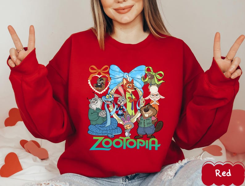 K&ouml;nnte beinhalten: Ein rotes Sweatshirt mit einer farbenfrohen Grafik von Charakteren aus dem Animationsfilm Zootopia. Das Design umfasst Judy Hopps, Nick Wilde und andere Charaktere, mit dem Wort "Zootopia" in gr&uuml;nen Buchstaben. Das Sweatshirt hat lange &Auml;rmel.