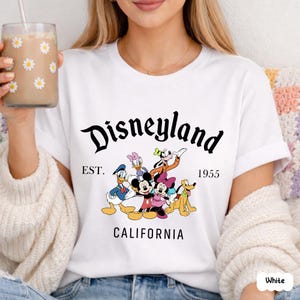 May include: White t-shirt featuring the word "Disneyland" in a black script font, with the text "EST. 1955 CALIFORNIA" below. The shirt has a graphic of Disney characters including Mickey Mouse, Minnie Mouse, Donald Duck, Daisy Duck, Goofy, and Pluto.