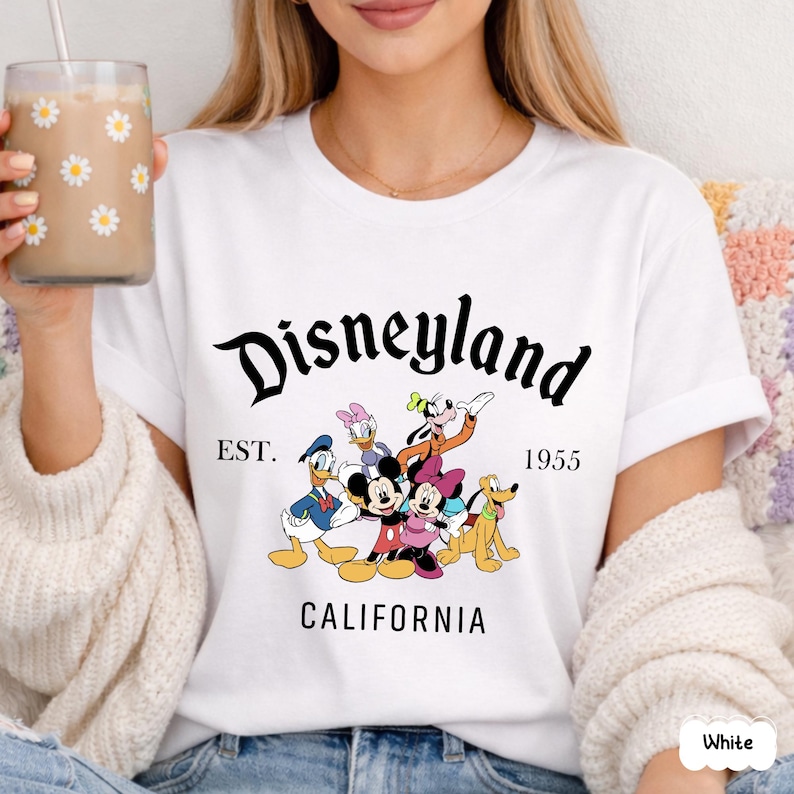 May include: White t-shirt featuring the word "Disneyland" in a black script font, with the text "EST. 1955 CALIFORNIA" below. The shirt has a graphic of Disney characters including Mickey Mouse, Minnie Mouse, Donald Duck, Daisy Duck, Goofy, and Pluto.