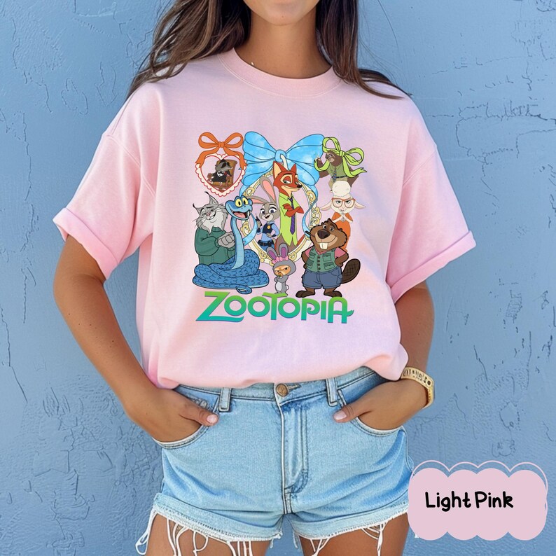 Może przedstawiać: Jasnor&oacute;żowa koszulka z kolorową grafiką postaci z filmu animowanego "Zwierzogr&oacute;d". Projekt zawiera niebieską kokardę, r&oacute;żne postacie zwierząt i słowo "Zootopia" w kolorze zielonym. Koszulka ma okrągły dekolt.