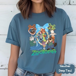 Może przedstawiać: T-shirt w kolorze głębokiego turkusu z kolorową grafiką postaci z animowanego filmu Zwierzogr&oacute;d. Projekt zawiera Judy Hopps, Nicka Wilde i inne postacie. Słowo "Zootopia" jest wydrukowane na dole w kolorze zielonym.