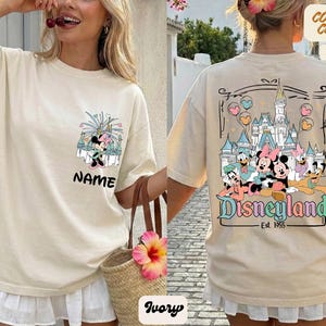 Puede incluir: Camiseta de color marfil con un gr&aacute;fico de Minnie Mouse y la palabra "NAME" en la parte delantera. La parte trasera muestra un gr&aacute;fico de Disneyland con personajes de dibujos animados y el texto "Disneyland Est. 1955".