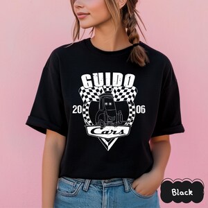 May include: Black t-shirt with a white graphic of the character Guido from the film Cars. The design includes the word "Guido", a chequered flag, the numbers "20" and "06", and the word "Cars".