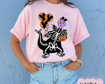 May include: Pale pink t-shirt with a black and white skeleton dragon design. The dragon holds Mickey Mouse balloons and a pumpkin basket. The dragon has purple and orange accents. The text "Light Pink" is at the bottom right.