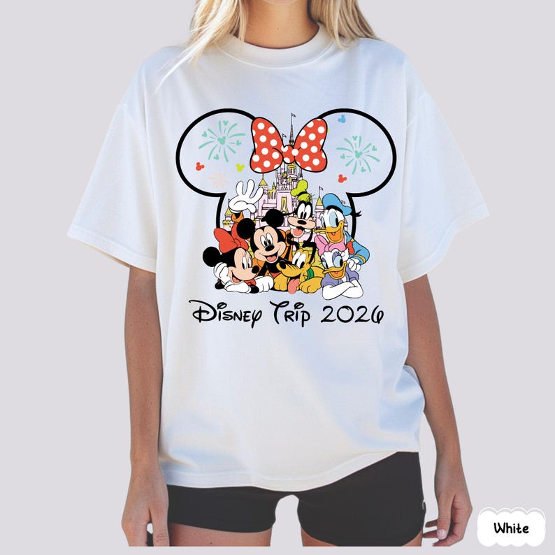 May include: White t-shirt featuring a graphic of Disney characters, including Mickey and Minnie Mouse, Goofy, Donald Duck, and Pluto. The design includes a castle, a polka-dot bow, and the text "Disney Trip 2026".