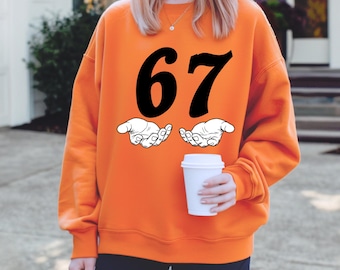 May include: Orange sweatshirt with the number 67 in large black font. Below the number are two white hands with open palms. The sweatshirt is a solid orange color.