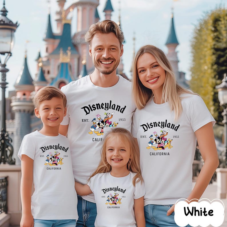 May include: A family of four wearing matching white t-shirts with the Disneyland logo. The shirts feature the text "Disneyland EST. 1955 CALIFORNIA" with cartoon characters. The family is smiling in front of a castle.
