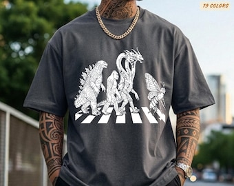 May include: Dark gray t-shirt featuring a white graphic of Godzilla, Bigfoot, a three-headed dragon, and a moth crossing a crosswalk. The shirt has the text "COMFORT COLORS" and "19 COLORS". The brand name "Pepper" is also visible.