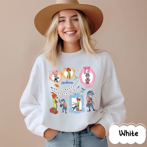 May include: White sweatshirt featuring colorful cartoon characters from the movie Zootopia. The design includes framed images of the characters, hearts, and the text "Zootopia" and "welcome to Zootopia".